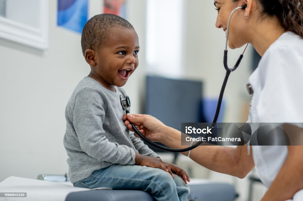 A young boy of African decent, sits up on an exam table as a female Paediatrician preforms a check-up on him.  The boy is dressed casually and smiling as the doctor listens to his heart.